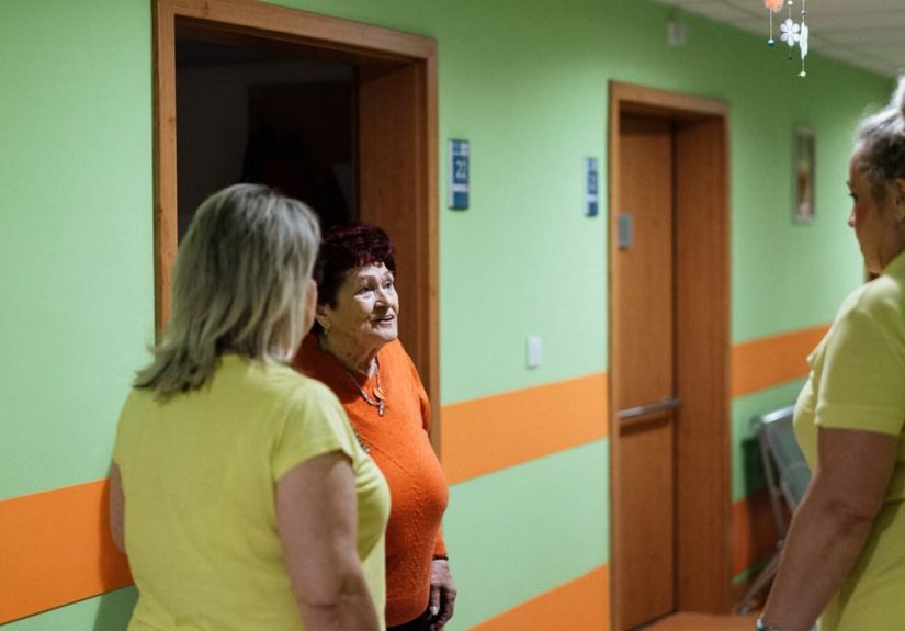 Senior woman engages in conversation with caregivers in a facility corridor.